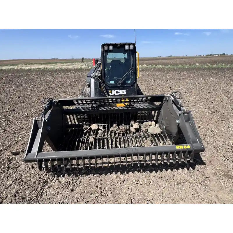 Front view of JCB skid steer with rock reel attachment lifting rocks from dry field soil under clear sky