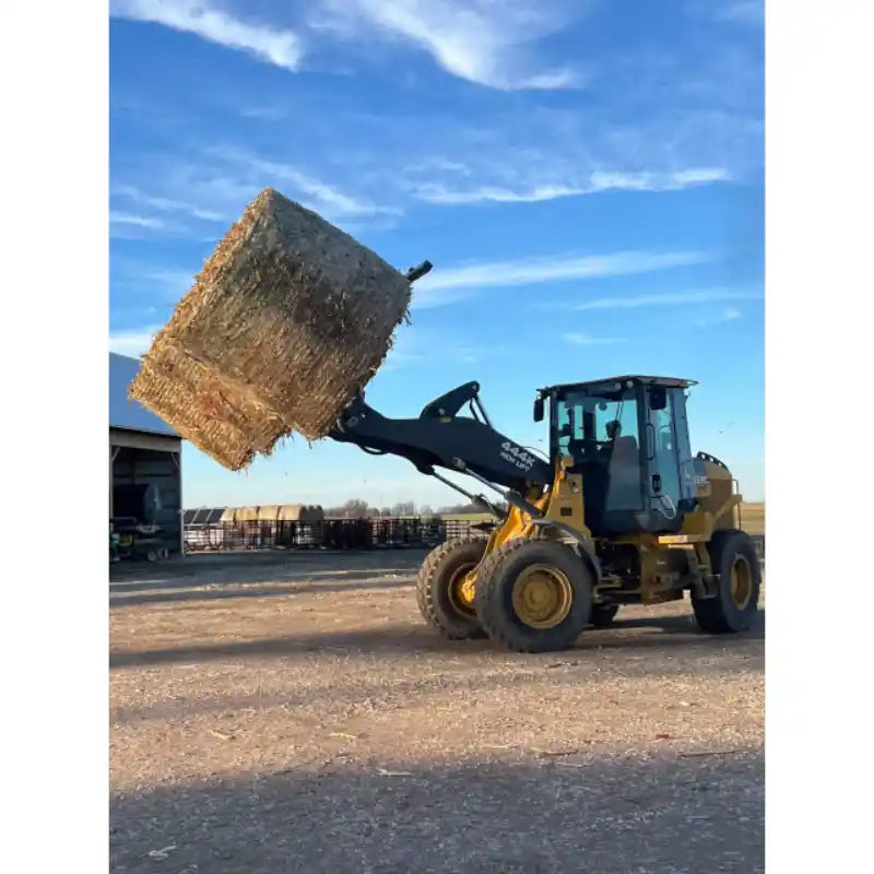 Yellow wheel loader lifting a large wrapped hay bale on dirt surface near farm buildings under clear sky