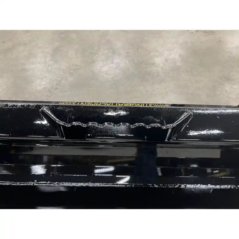 Top view of black steel skid steer material bucket edge with reinforced grip and weld details on concrete floor