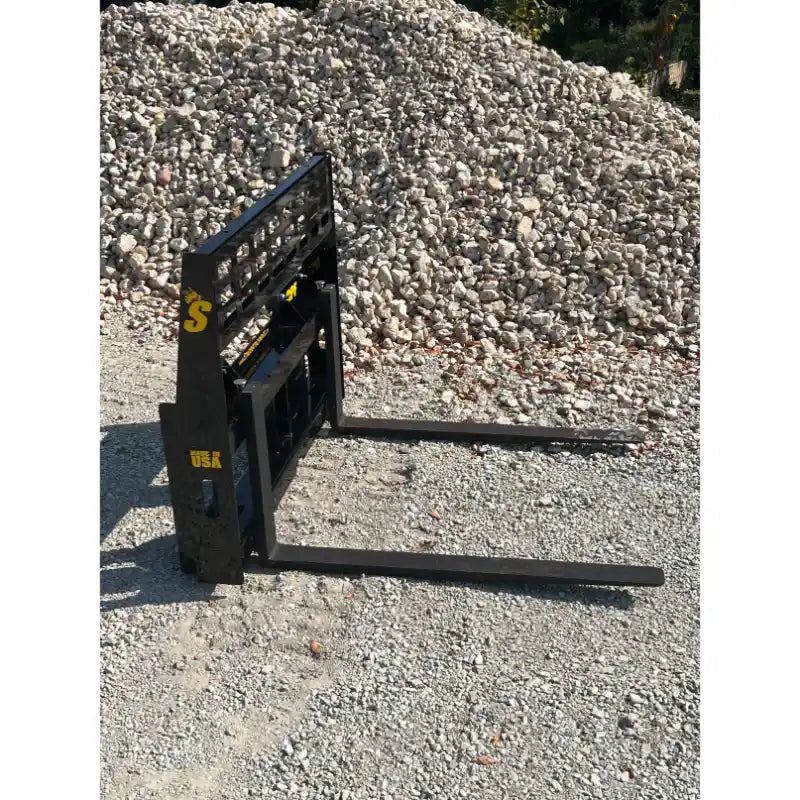 Black steel skid steer pallet fork attachment with grid backrest and two extended forks on gravel surface outdoors