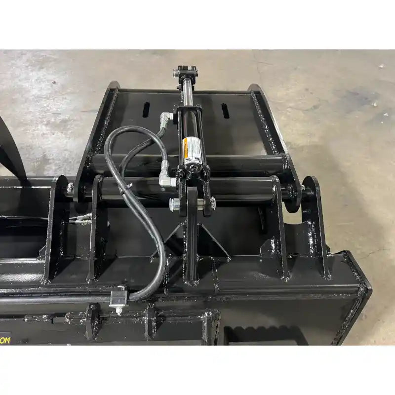 Close-up of black metal grapple attachment with hydraulic cylinder and hoses on concrete floor indoors