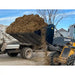 Skid steer loader bucket dumping organic mulch into a farm trailer at an outdoor rural site