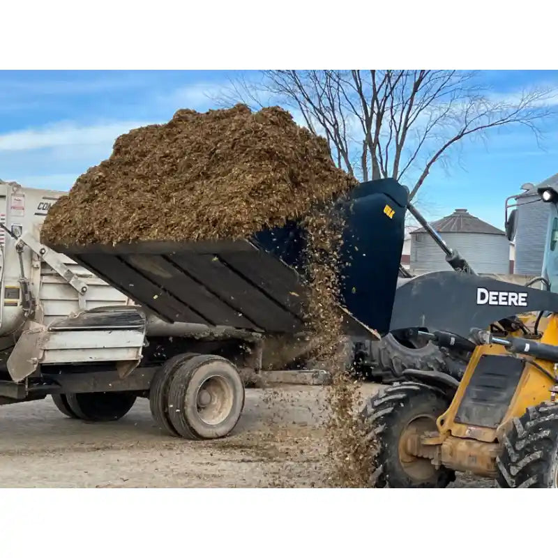 Skid steer loader bucket dumping organic mulch into a farm trailer at an outdoor rural site