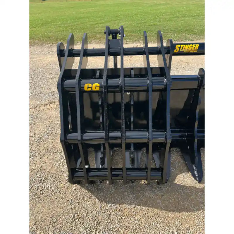 Black steel grapple attachment with multiple vertical tines and reinforced crossbars resting on gravel outdoors