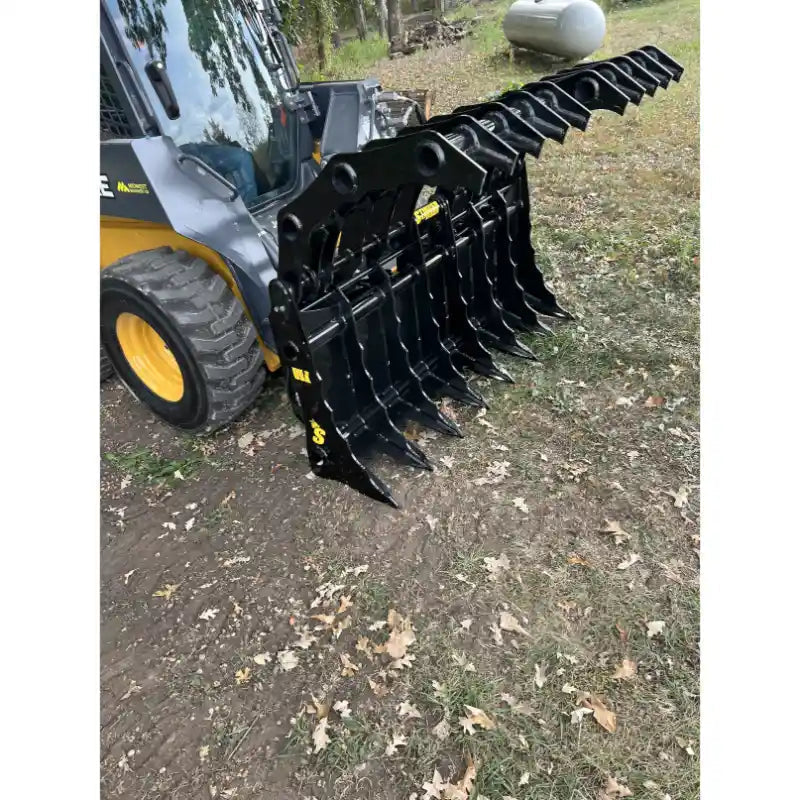 Skid steer loader with extended black steel grapple attachment featuring multiple curved tines on dirt and grass surface outdoors