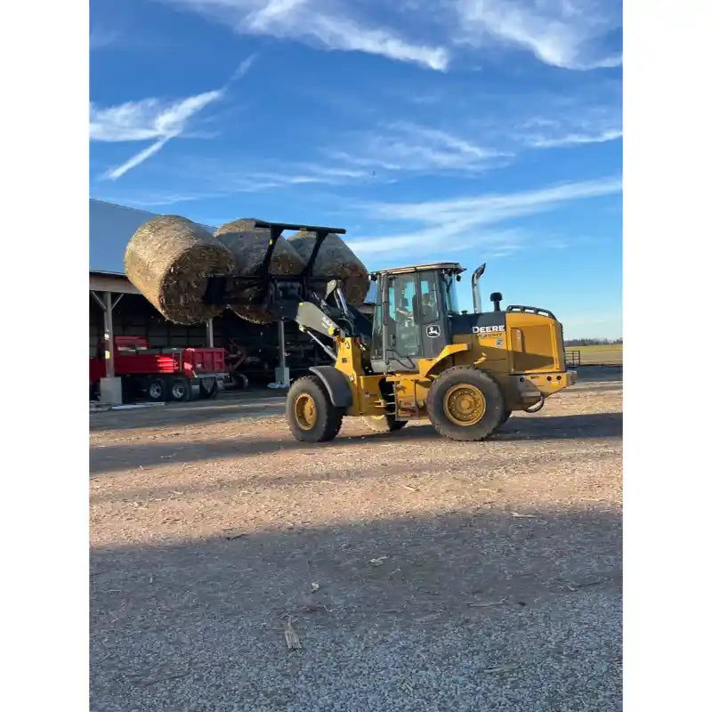 Yellow wheel loader lifting two large round hay bales on a dirt surface near farm structures under blue sky
