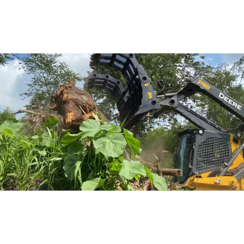 Yellow Deere skid steer with black clam grapple attachment lifting uprooted tree among dense green vegetation under blue sky