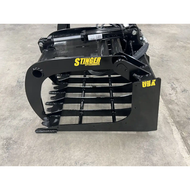 Black steel skid steer grapple attachment with thick spaced tines, hydraulic cylinder, and reinforced side plate on concrete floor