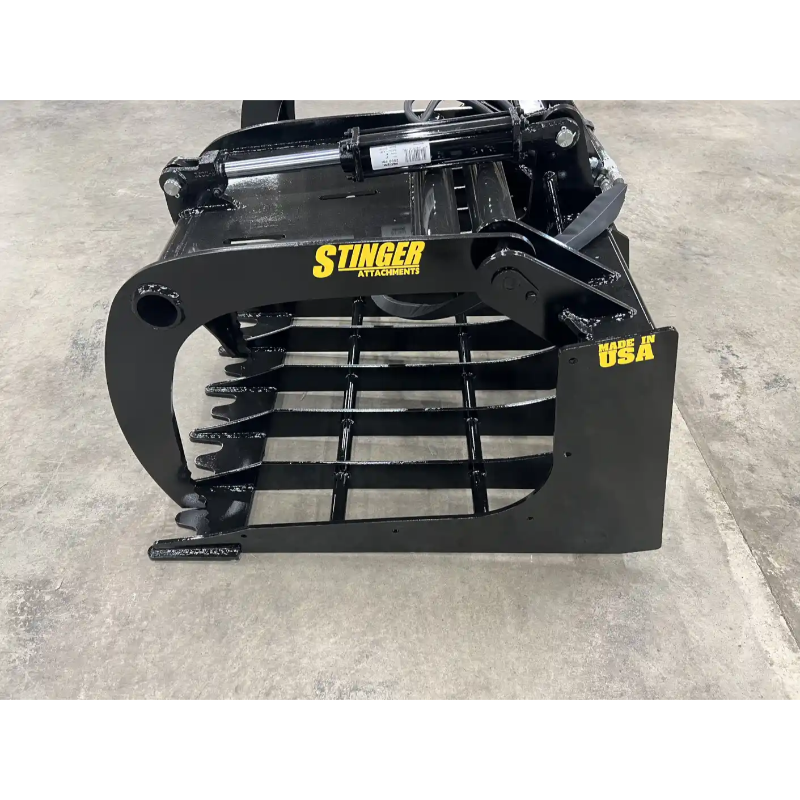 Black steel skid steer grapple attachment with thick spaced tines, hydraulic cylinder, and reinforced side plate on concrete floor