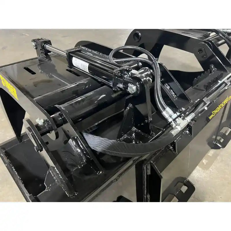 Black steel demolition grapple attachment focusing on hydraulic cylinder and hose connections indoors on concrete floor