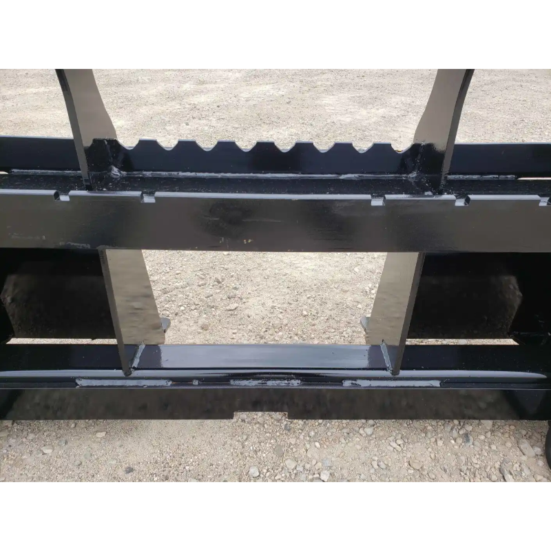 Close-up of black painted steel skid steer pallet fork attachment with reinforced tines and mounting frame on gravel surface