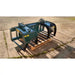 Green-painted steel skid steer grapple attachment with two hydraulic cylinders and curved tines on wooden pallet outdoors