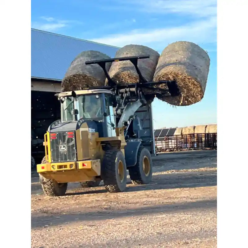 Loader with front attachment carrying three large round straw bales on dirt surface near farm buildings