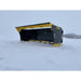 Black and yellow skid steer snow pusher attachment resting on snow-covered ground outdoors with visible cutting edge bolts