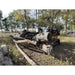 Tracked skid steer loader with black grapple attachment carrying large tree logs outdoors in wooded area