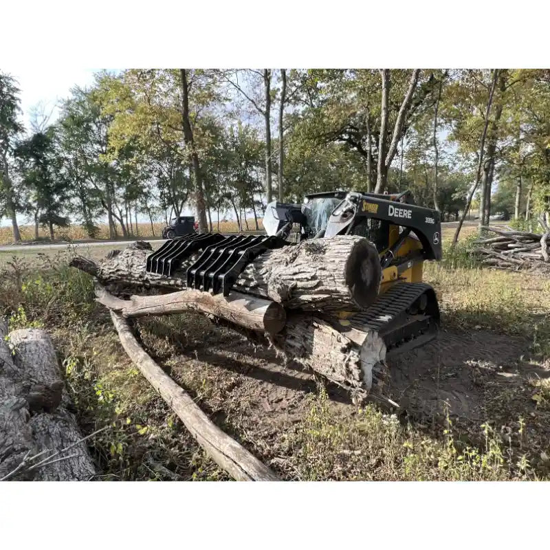 Tracked skid steer loader with black grapple attachment carrying large tree logs outdoors in wooded area