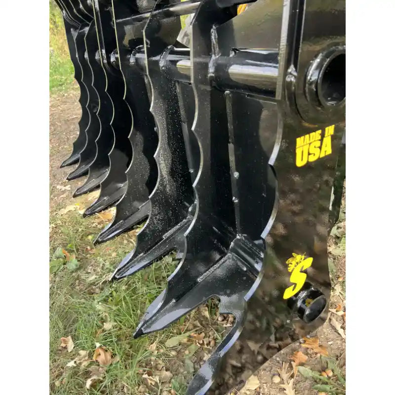 Close-up view of black steel grapple attachment teeth with yellow "Made in USA" and logo markings outdoors on grass and dirt