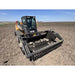 Front angled view of tracked skid steer with rock reel attachment clearing rocky soil in open field under blue sky