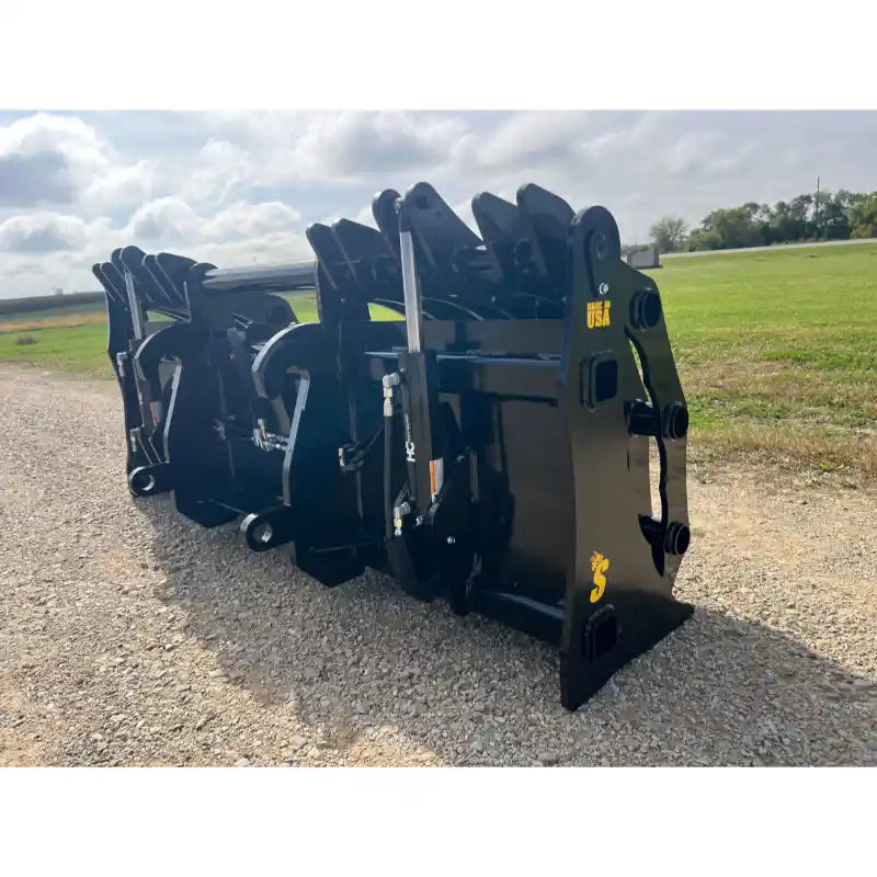Row of black steel clam grapple attachments with hydraulic components resting on gravel outdoors under cloudy sky