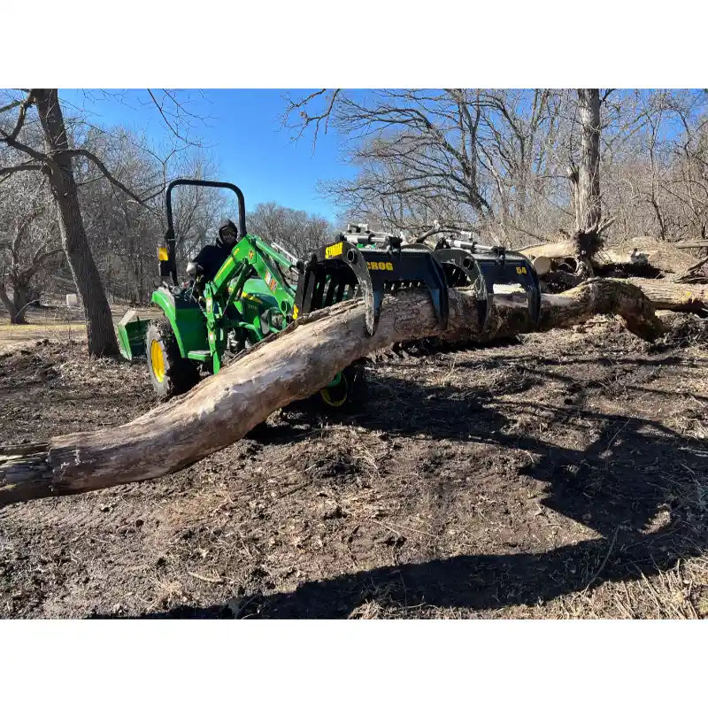 Green compact tractor with a black grapple attachment lifting a large tree log over bare ground with leafless trees nearby