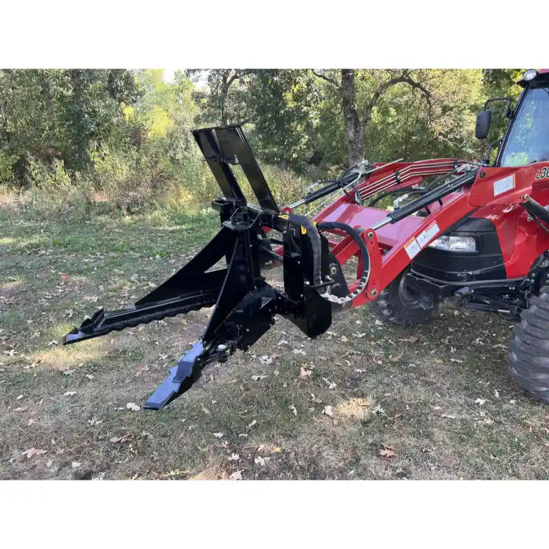 Black steel tractor loader attachment with serrated gripping arms and hydraulic hoses on grassy ground with trees