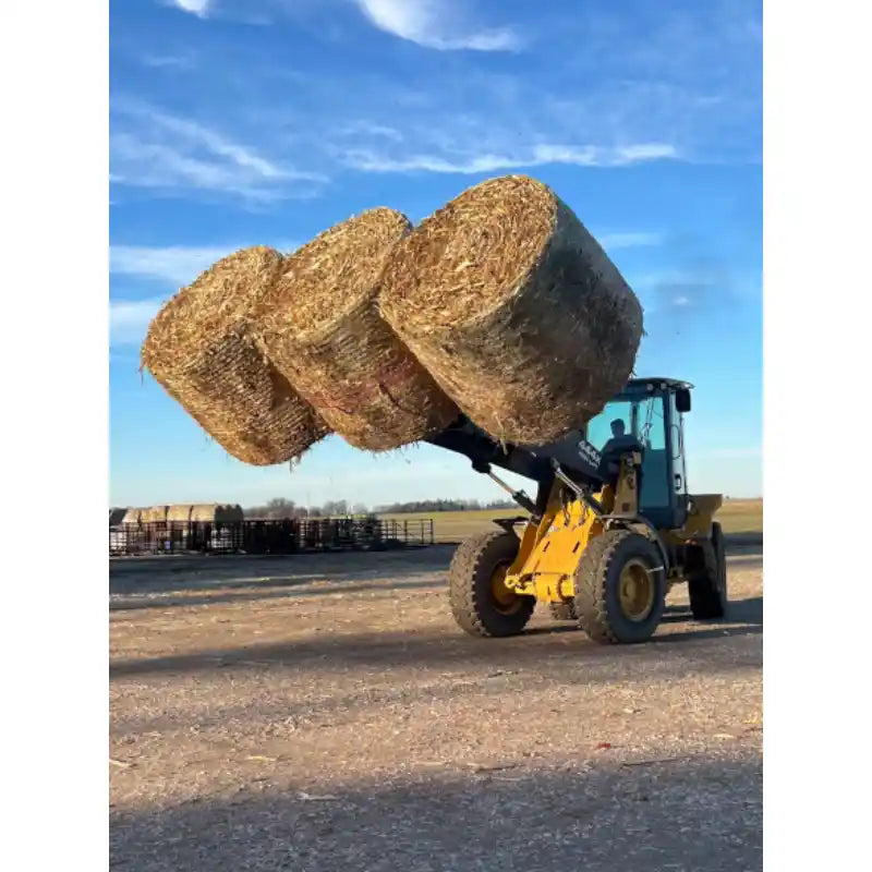 Yellow skid steer loader lifting three large round straw bales outdoors on dirt surface under clear sky