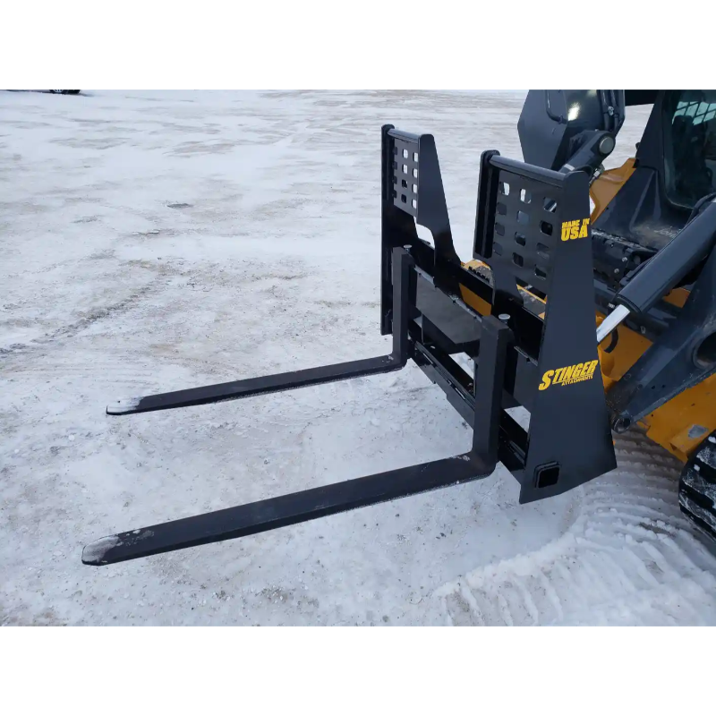 Black painted steel skid steer pallet fork attachment with wide fork tines and reinforced mounting brackets on snow-covered ground