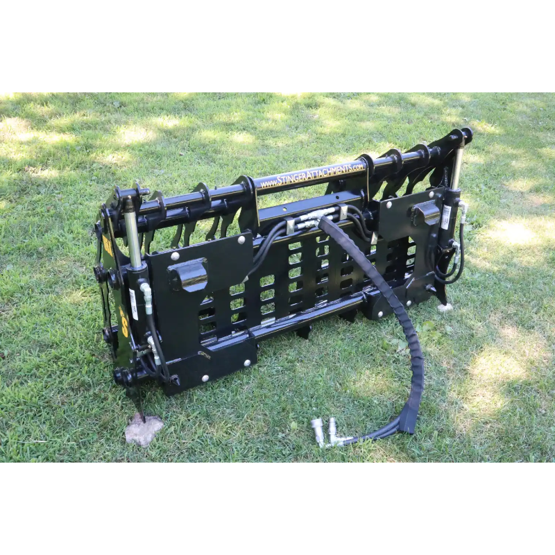 Black compact clam grapple attachment with hydraulic hoses resting on grass outdoors with sunlight and shadow patterns