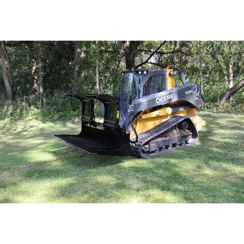 Compact track loader with black open rock grapple attachment and Deere branding on a grassy clearing near trees