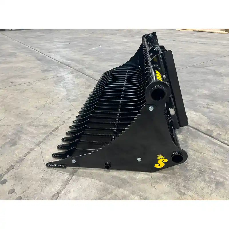 Black metal rock bucket skid steer attachment with curved spaced tines and reinforced mounting points on concrete floor