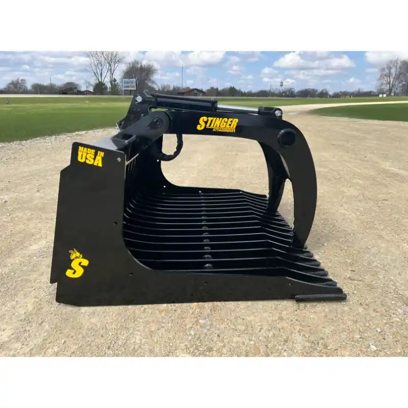 Black steel rock grapple attachment with spaced flat tines, hydraulic pivot arm, and yellow Stinger logo on gravel surface outdoors
