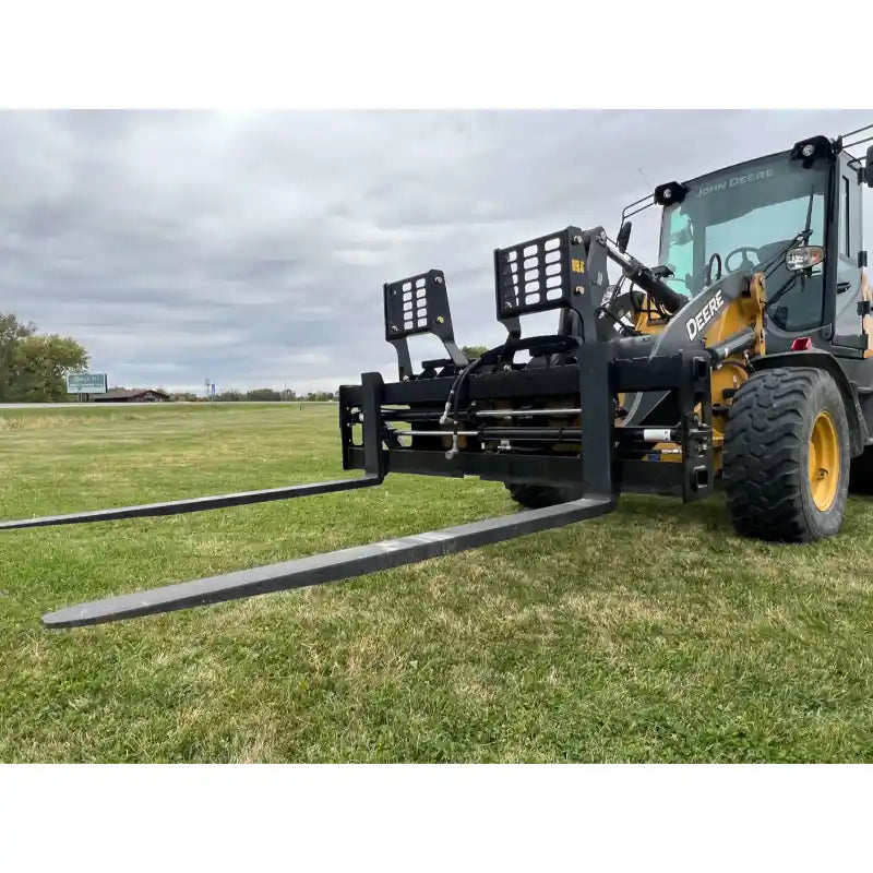 Yellow Deere skid steer loader fitted with black pallet fork attachment and extended fork tines on mowed grassy field under cloudy sky