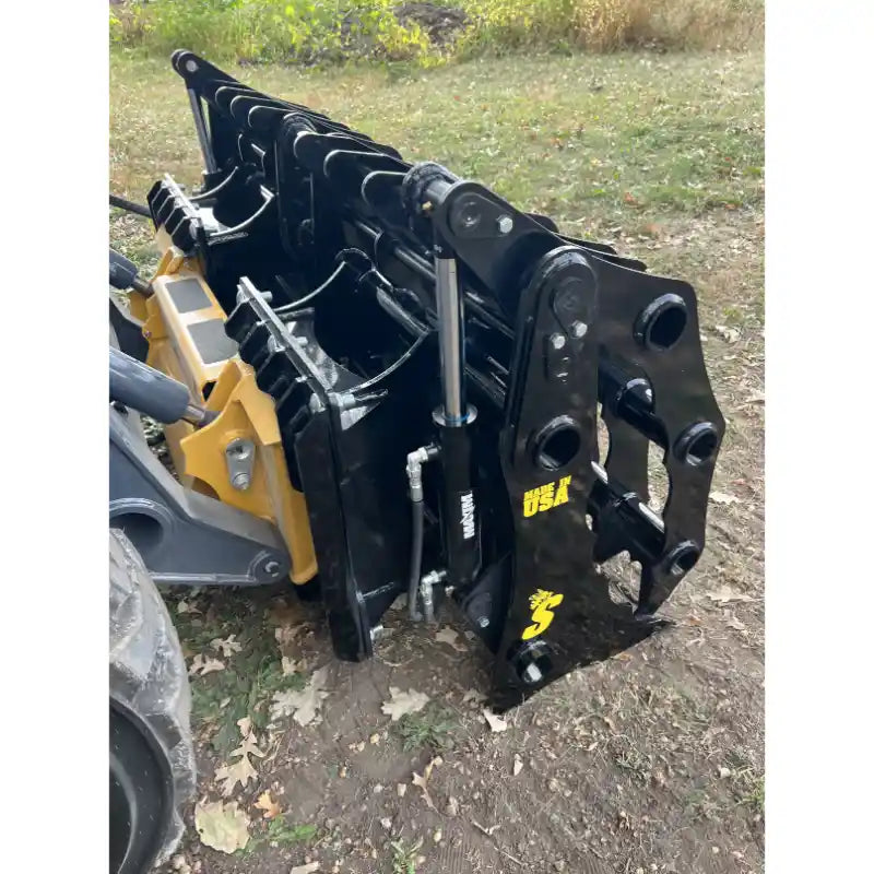 Black steel clam grapple attachment with hydraulic cylinders and mounting points resting on dirt and leaves outdoors