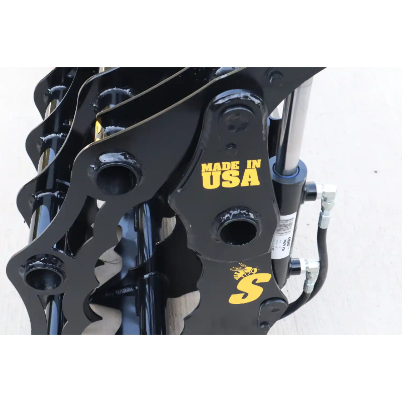 Close-up of black steel grapple attachment with visible hydraulic hoses and yellow "Made in USA" marking on light surface