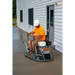 Operator wearing white hard hat and orange shirt seated on a ride-on power trowel smoothing fresh concrete near building siding
