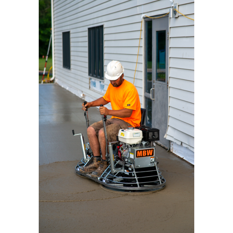 Operator wearing white hard hat and orange shirt seated on a ride-on power trowel smoothing fresh concrete near building siding