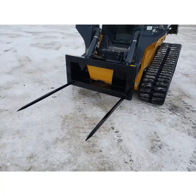 Tracked skid steer with black bale spear attachment featuring two long pointed steel spikes on ground