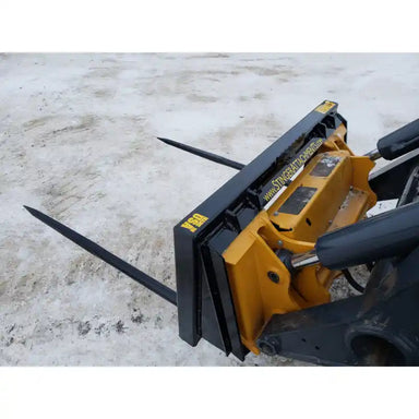 Yellow and black skid steer bale spear attachment with two long pointed steel spears on concrete surface