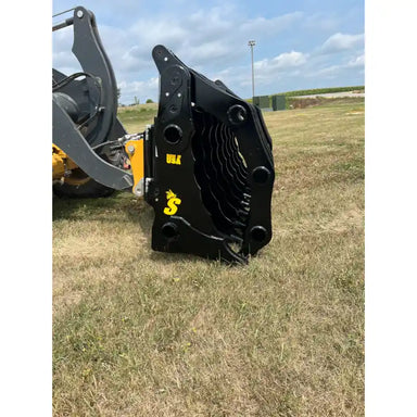 Black steel clam grapple attachment with multiple tines mounted on loader arm over grassy field under partly cloudy sky