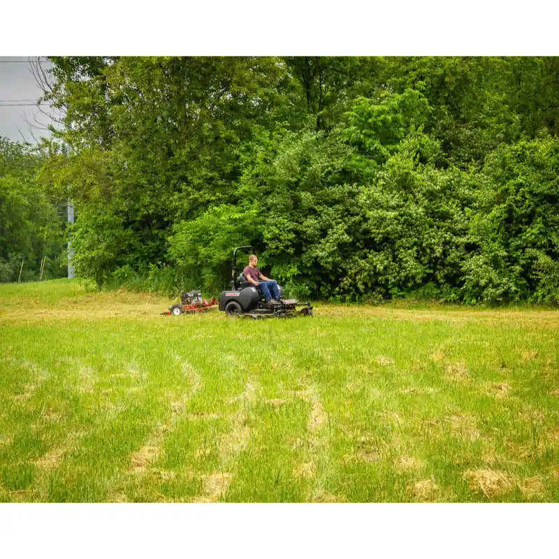 Operator using a zero-turn mower trimming a grassy field near dense trees, mower positioned center-right in the frame, gallery position 1