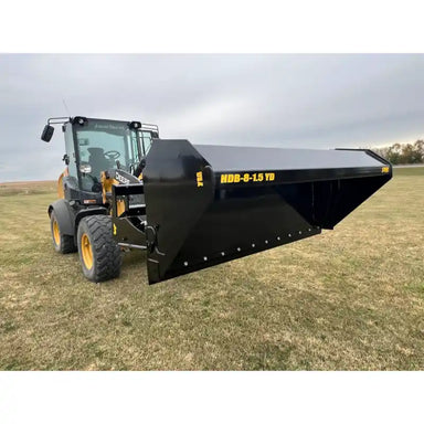 Black steel hydraulic dump bucket attachment mounted on loader with visible yellow lettering on grassy field
