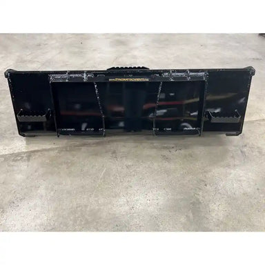 Black steel skid steer attachment with reinforced mounting points and smooth surface resting on concrete floor indoors