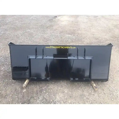 Rear view of a black steel skid steer material bucket attachment with mounting plates resting on dirt surface