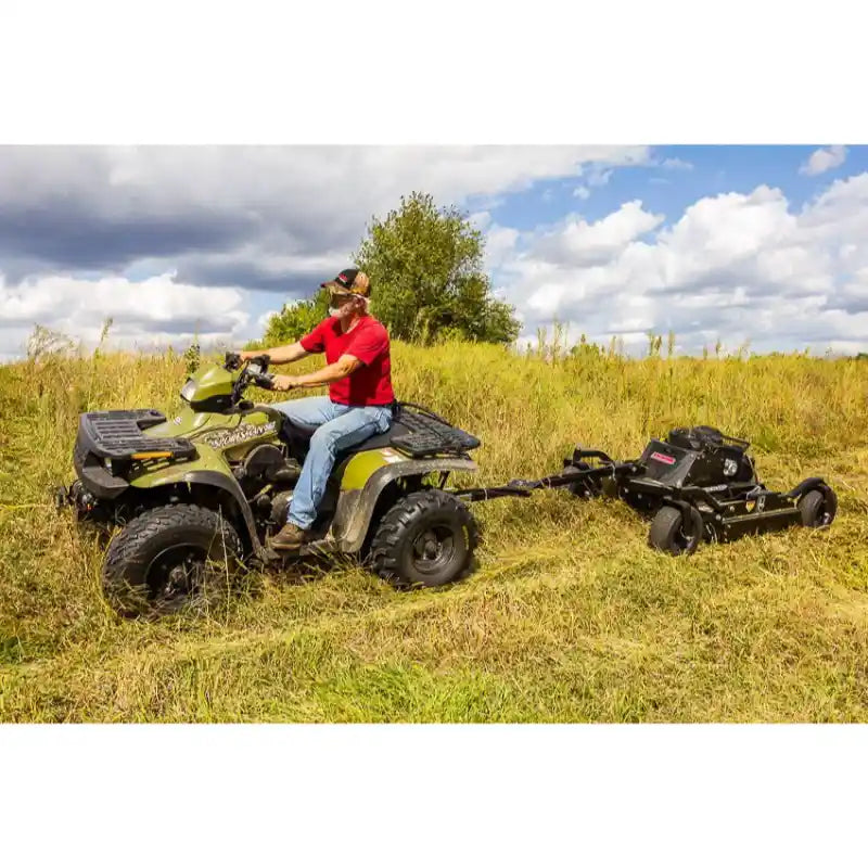 Operator on ATV pulling Swisher 44" brush cutter with 14.5HP Kawasaki engine across rough grassy terrain, gallery position 1