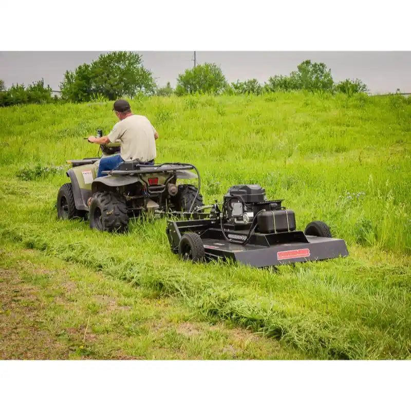 A person operates an ATV pulling a Swisher 44 Rough Cut Mower – 14.5HP 12V Country Cut, effectively mowing tall grass in a field with trees in the background.