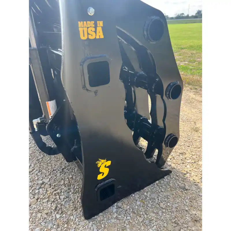 Close-up of black steel grapple attachment with hydraulic fittings and yellow "Made in USA" and logo markings on gravel outdoors