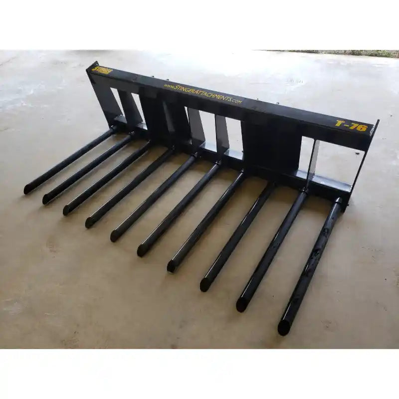 Skid steer attachment with nine long black metal tines and rectangular mounting frame on concrete floor indoors