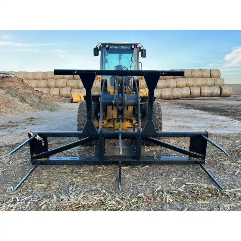 Black steel bale spear skid steer attachment with five pointed tines mounted on yellow loader outdoors on dirt surface