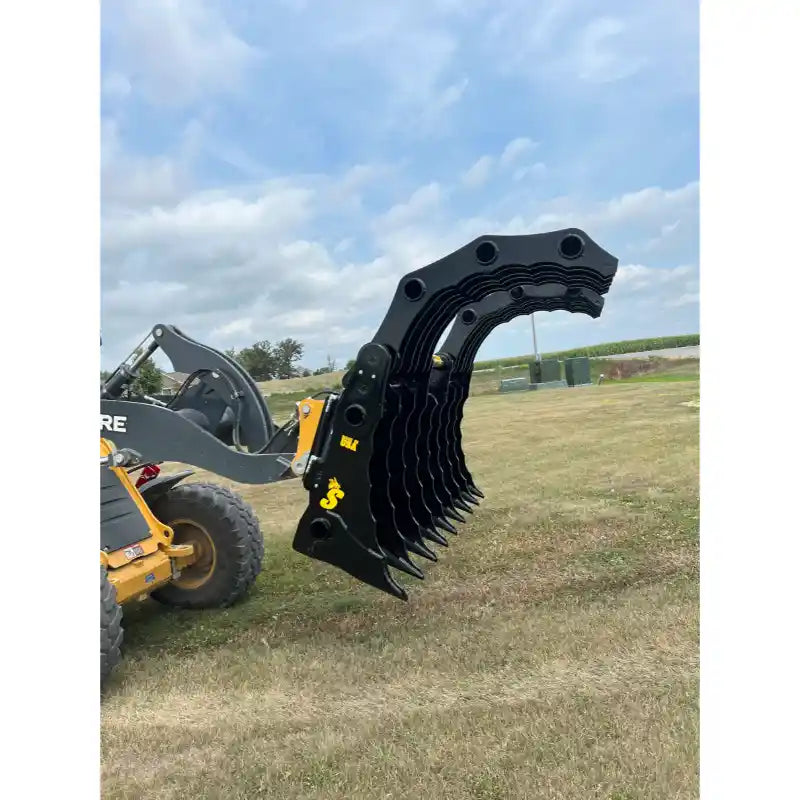 Yellow loader with black multi-tine clam-style grapple attachment raised over grassy field under partly cloudy sky