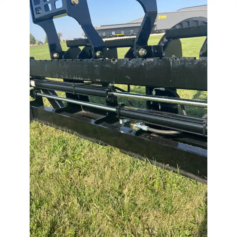 Close-up of black hydraulic skid steer attachment with metal bars and bolts resting on grass outdoors